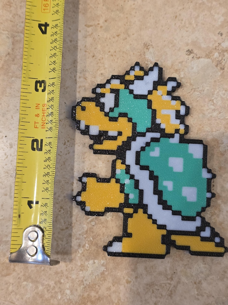 Puede incluir: Una figura pixelada de Bowser, de aproximadamente 7,6 cm de altura, con caparaz&oacute;n verde, vientre y cabeza amarillos y detalles blancos. La figura tiene un acabado brillante y est&aacute; delineada en negro. Una cinta m&eacute;trica amarilla est&aacute; en la imagen.