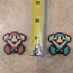 Puede incluir: Dos figuras de pixel art, una roja y otra turquesa, junto a una cinta m&eacute;trica. Las figuras miden aproximadamente 2,5 cm de alto. Las figuras est&aacute;n hechas de peque&ntilde;as cuentas de colores.