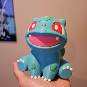 Peut inclure: Figurine bleue de Bulbasaur avec des taches vertes et une bouche rose. La figurine a des dents blanches et des yeux rouges. Bulbasaur est assis et a des orteils blancs.