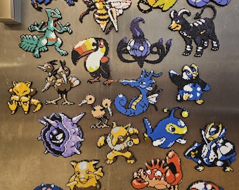 May include: A collection of colorful pixel art magnets featuring various Pokémon characters. The magnets are made from small, fused beads and include a variety of creatures in different shapes and colors, such as a toucan, a bee, and a blue octopus.