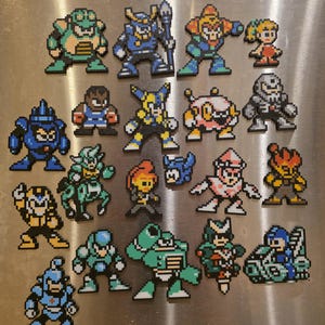 Könnte beinhalten: Eine Sammlung von Pixel-Art-Magneten mit Charakteren aus der Videospielserie Mega Man. Die Magnete zeigen verschiedene Charaktere in leuchtenden Farben, darunter Blau, Grün, Orange und Gelb. Jeder Magnet hat ein einzigartiges Design.