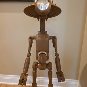 Op de afbeelding: Een bruine, gelede robotfiguur met een grote, cirkelvormige lens op zijn hoofd. De robot heeft een slanke nek, een rechthoekige romp en gelede ledematen. Hij staat op een houten vloer, met een muur en kunstwerk op de achtergrond.