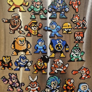 May include: A collection of colorful pixel art magnets featuring characters from the Mega Man video game series. The magnets are in various colors, including blue, orange, green, and yellow, and are arranged on a metallic surface.