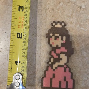 Puede incluir: Figura pixelada de la Princesa Peach, de aproximadamente 5 cm de altura, con un vestido rosa y cabello casta&ntilde;o, con una peque&ntilde;a corona. La figura est&aacute; junto a una cinta m&eacute;trica amarilla.