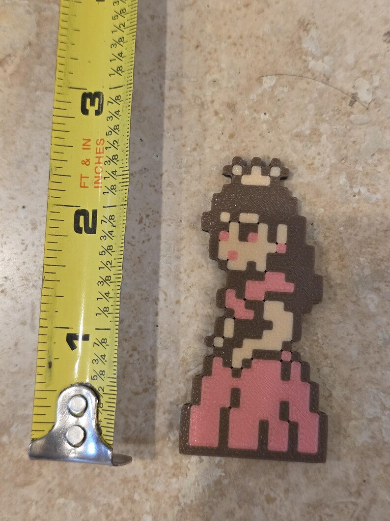 Puede incluir: Figura pixelada de la Princesa Peach, de aproximadamente 5 cm de altura, con un vestido rosa y cabello casta&ntilde;o, con una peque&ntilde;a corona. La figura est&aacute; junto a una cinta m&eacute;trica amarilla.