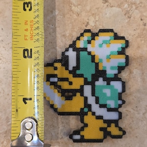 Puede incluir: Una figura pixelada de un personaje de videojuego, principalmente amarillo, blanco y verde, se muestra junto a una cinta m&eacute;trica. La figura mide aproximadamente 5 cm de alto. El personaje es un dise&ntilde;o retro, que recuerda a los videojuegos cl&aacute;sicos de 8 bits.