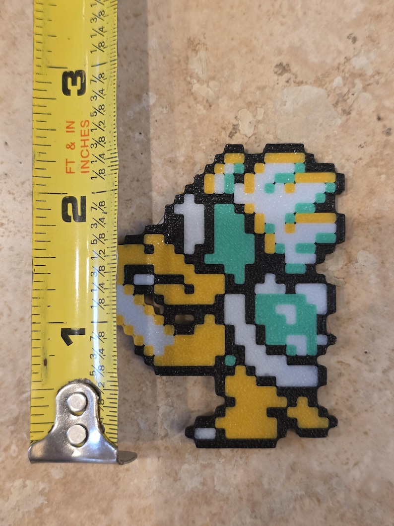 Puede incluir: Una figura pixelada de un personaje de videojuego, principalmente amarillo, blanco y verde, se muestra junto a una cinta m&eacute;trica. La figura mide aproximadamente 5 cm de alto. El personaje es un dise&ntilde;o retro, que recuerda a los videojuegos cl&aacute;sicos de 8 bits.