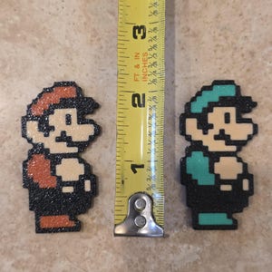 Puede incluir: Figuras pixeladas de Mario y Luigi, cada una de aproximadamente 5 cm de altura, se muestran juntas. Mario tiene un sombrero y un mono rojos, mientras que Luigi lleva un sombrero y un mono verdes. Una cinta m&eacute;trica amarilla est&aacute; entre ellos.