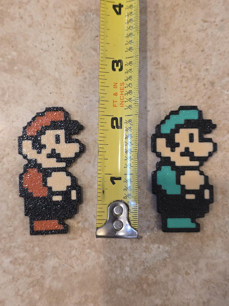 Puede incluir: Figuras pixeladas de Mario y Luigi, cada una de aproximadamente 5 cm de altura, se muestran juntas. Mario tiene un sombrero y un mono rojos, mientras que Luigi lleva un sombrero y un mono verdes. Una cinta m&eacute;trica amarilla est&aacute; entre ellos.