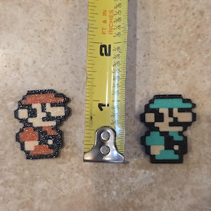 Puede incluir: Dos figuras de personajes de pixel art, una en rojo y naranja, la otra en verde azulado y azul. Las figuras miden aproximadamente 2,5 cm de alto. Las figuras est&aacute;n sobre una superficie marr&oacute;n claro.