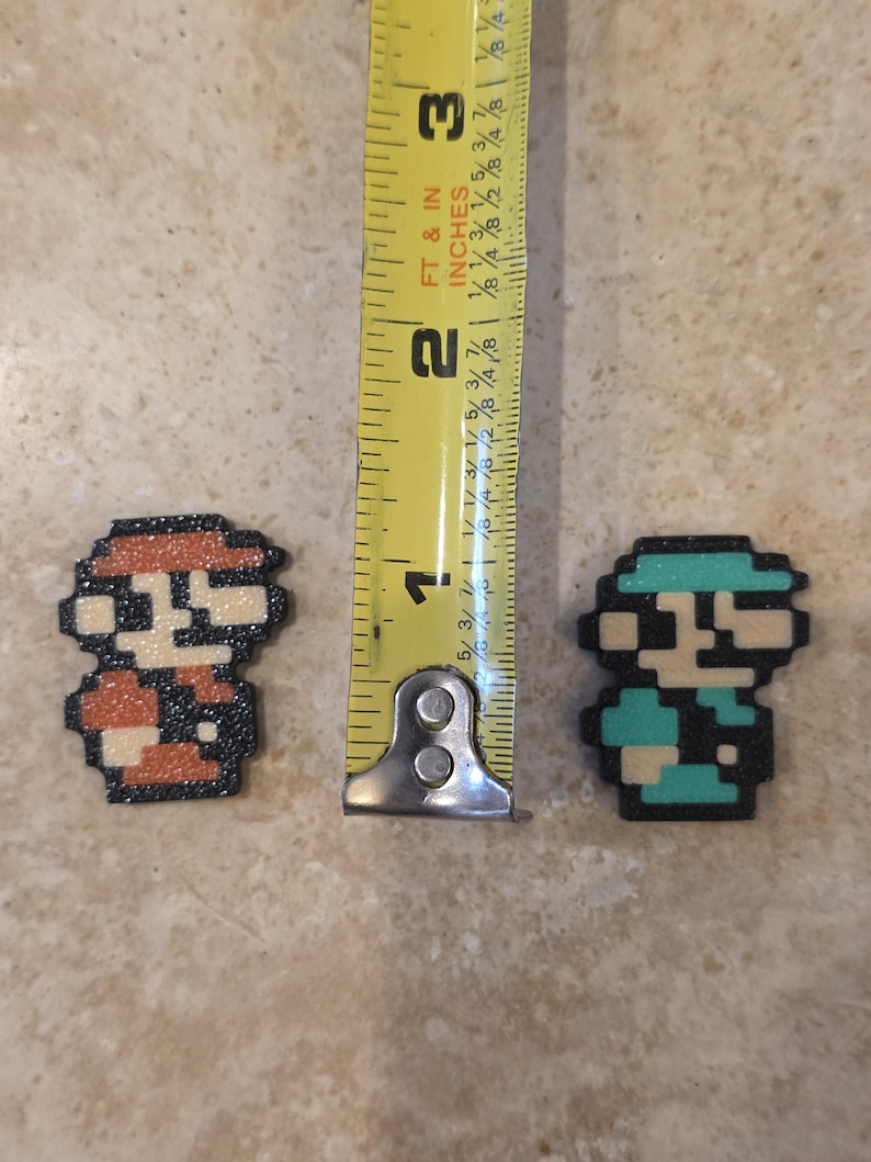 Puede incluir: Dos figuras de personajes de pixel art, una en rojo y naranja, la otra en verde azulado y azul. Las figuras miden aproximadamente 2,5 cm de alto. Las figuras est&aacute;n sobre una superficie marr&oacute;n claro.