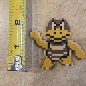 Puede incluir: Una figura pixelada de Koopa Troopa de la serie de videojuegos Super Mario Bros. La figura es amarilla, marr&oacute;n y beige, y mide aproximadamente 6,35 cm de altura. La figura est&aacute; sobre una superficie de color neutro.