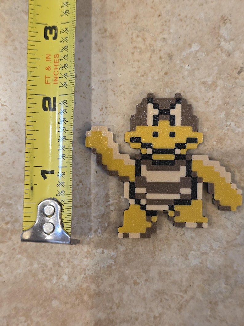 Puede incluir: Una figura pixelada de Koopa Troopa de la serie de videojuegos Super Mario Bros. La figura es amarilla, marr&oacute;n y beige, y mide aproximadamente 6,35 cm de altura. La figura est&aacute; sobre una superficie de color neutro.