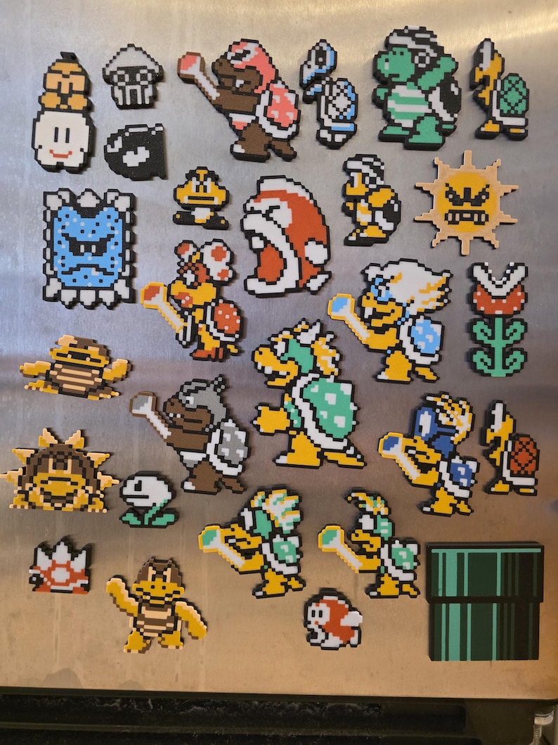 Puede incluir: Una colecci&oacute;n de imanes pixelados con personajes de la serie de videojuegos Super Mario. Los imanes incluyen a Mario, Bowser, Goombas y Koopa Troopas, en colores vibrantes. La colecci&oacute;n incluye una tuber&iacute;a verde.