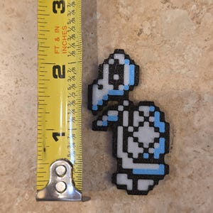 Puede incluir: Una figura de tortuga pixelada en negro, blanco y azul claro, de aproximadamente 6,35 cm de altura. La figura est&aacute; junto a una cinta m&eacute;trica amarilla, que muestra su tama&ntilde;o. La tortuga tiene un caparaz&oacute;n y una cabeza, que recuerda a un personaje de videojuego.