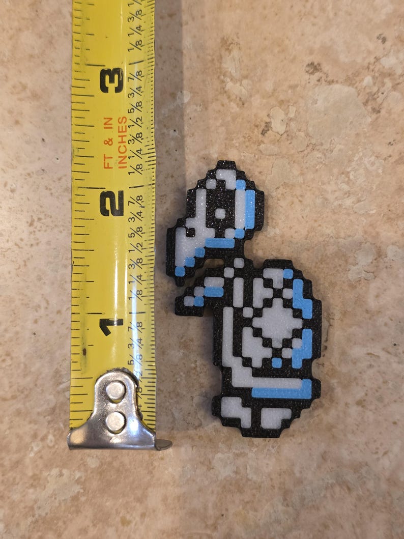 Puede incluir: Una figura de tortuga pixelada en negro, blanco y azul claro, de aproximadamente 6,35 cm de altura. La figura est&aacute; junto a una cinta m&eacute;trica amarilla, que muestra su tama&ntilde;o. La tortuga tiene un caparaz&oacute;n y una cabeza, que recuerda a un personaje de videojuego.