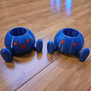 May include: Two blue, spherical planters with red eyes and smiling faces. Each planter has two blue, oval-shaped feet. The planters are made of a 3D-printed material and are sitting on a wooden surface.