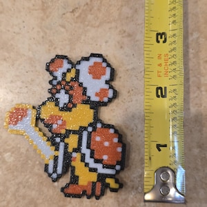 Puede incluir: Personaje pixelado de la serie de videojuegos Super Mario Bros. La figura mide aproximadamente 7,6 cm de alto, con un cuerpo amarillo, un caparaz&oacute;n naranja y un gorro de hongo blanco. El personaje est&aacute; hecho de peque&ntilde;as cuentas de colores.