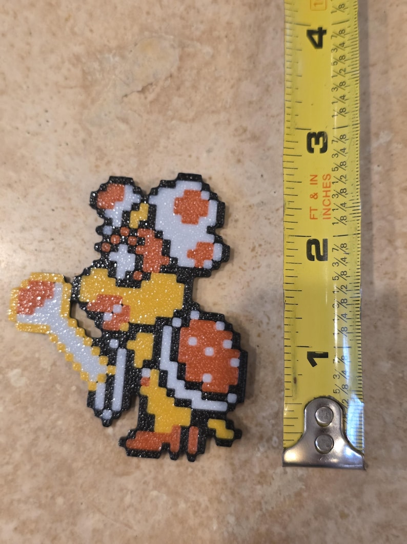 Puede incluir: Personaje pixelado de la serie de videojuegos Super Mario Bros. La figura mide aproximadamente 7,6 cm de alto, con un cuerpo amarillo, un caparaz&oacute;n naranja y un gorro de hongo blanco. El personaje est&aacute; hecho de peque&ntilde;as cuentas de colores.