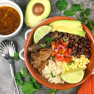 May include: A colorful bowl of food with various ingredients, including black beans, corn, rice, and avocado slices. A small white bowl of soup and a silver spoon are next to the bowl. A lime wedge adds a fresh touch.