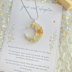 Pode incluir: Um colar com pingente de meia-lua envolto em fio prateado, decorado com pequenos cristais verdes e amarelo-dourados. O colar é exibido sobre um cartão branco com o texto "your next chapter..." e uma mensagem inspiradora sobre novos começos.