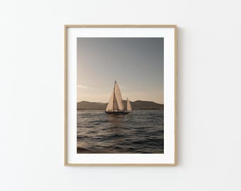 May include: A framed print featuring a sailboat on the water. The boat has white sails and is sailing on a body of water with mountains in the background. The sky is a light blue color.