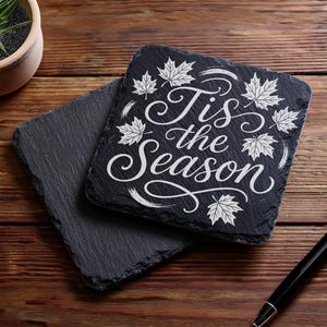 May include: Two square black slate coasters with the text "Tis the Season" in white script and maple leaf designs. One coaster is stacked on top of the other. A black pen rests on a wooden surface.