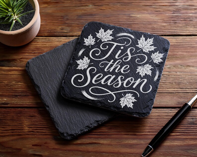 May include: Two square black slate coasters with the text "Tis the Season" in white script and maple leaf designs. One coaster is stacked on top of the other. A black pen rests on a wooden surface.