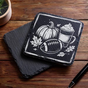 May include: Black slate coasters with a white graphic of a pumpkin, football, coffee cup, and leaves. One coaster is stacked on top of another. A pen is in the bottom right corner. The coasters are on a wooden surface.