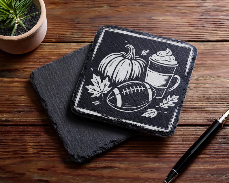 May include: Black slate coasters with a white graphic of a pumpkin, football, coffee cup, and leaves. One coaster is stacked on top of another. A pen is in the bottom right corner. The coasters are on a wooden surface.