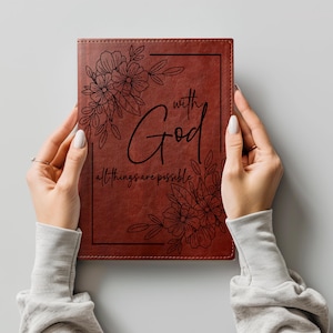 May include: A rectangular brown leather-like journal featuring a black engraved floral border design. The center text reads "with God all things are possible" in a combination of script and serif fonts, framed by black line art flowers.