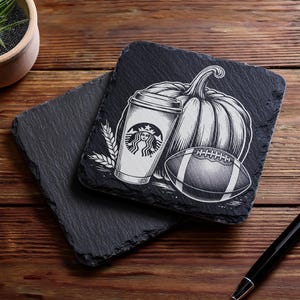 May include: Two black slate coasters with a fall-themed design. One coaster features a Starbucks coffee cup, a pumpkin, and a football. The other coaster is plain. The coasters are on a wooden surface.