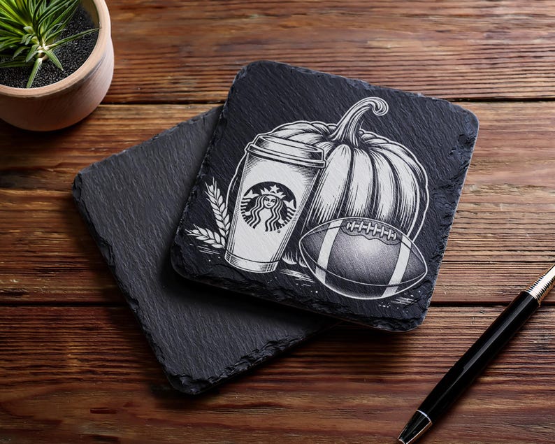 May include: Two black slate coasters with a fall-themed design. One coaster features a Starbucks coffee cup, a pumpkin, and a football. The other coaster is plain. The coasters are on a wooden surface.