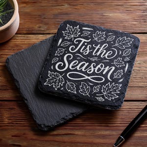 May include: Two square, dark gray slate coasters. One coaster has the phrase "Tis the Season" in white script with leaf illustrations. The coasters are on a wooden surface with a pen and a small potted plant.