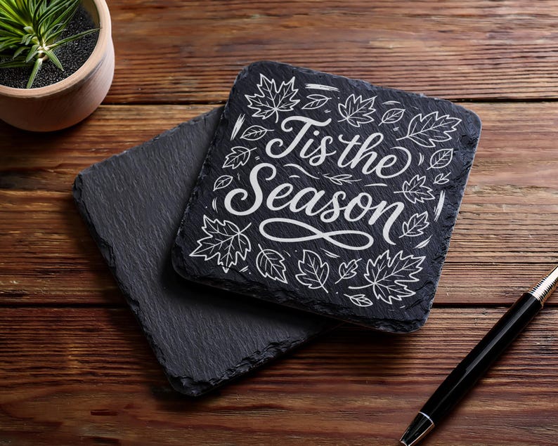 May include: Two square, dark gray slate coasters. One coaster has the phrase "Tis the Season" in white script with leaf illustrations. The coasters are on a wooden surface with a pen and a small potted plant.