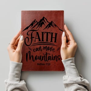 May include: A brown leather-textured journal featuring a black graphic of mountain peaks at the top. The text reads "FAITH can move Mountains" in a mix of serif and script fonts, followed by the citation "Matthew 17:20" in a simple font.