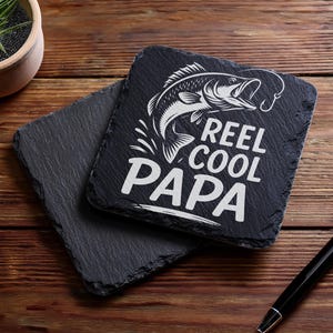 May include: Two square black slate coasters with a rough, natural edge. One coaster features a white graphic of a fish and the words "REEL COOL PAPA". The coasters are on a wooden surface.