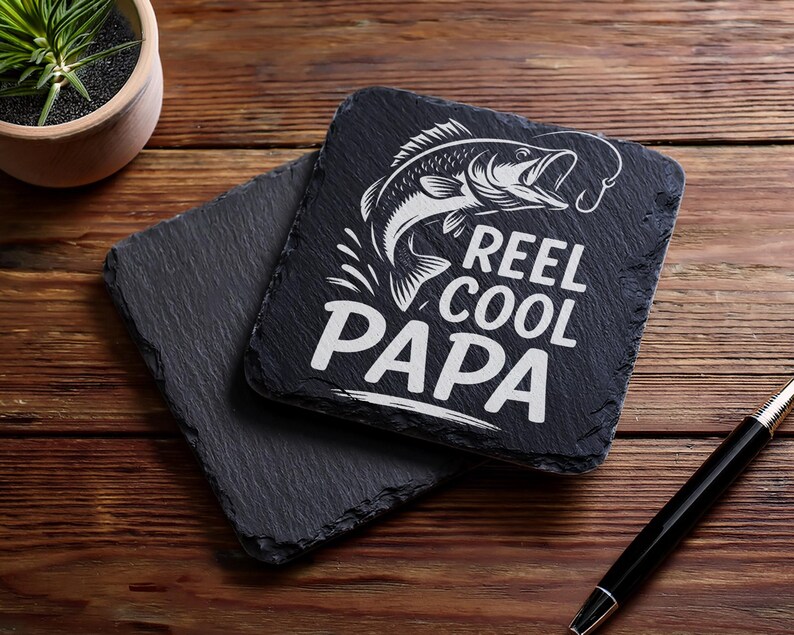 May include: Two square black slate coasters with a rough, natural edge. One coaster features a white graphic of a fish and the words "REEL COOL PAPA". The coasters are on a wooden surface.
