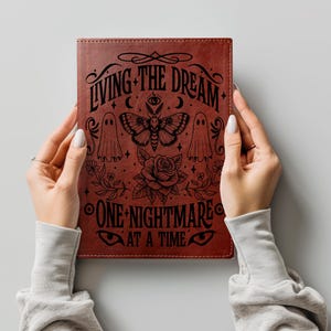 May include: A rectangular dark brown notebook featuring black line art of a moth, two ghosts, a rose, and an eye. The text reads: LIVING THE DREAM ONE NIGHTMARE AT A TIME. The cover has a textured finish with visible stitching along the edges.