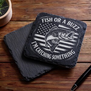 May include: Two square black slate coasters. One coaster has the text "FISH OR A BUZZ I'M CATCHING SOMETHING" with a fish graphic and an American flag design. The other coaster is plain. A pen is on the wooden surface.