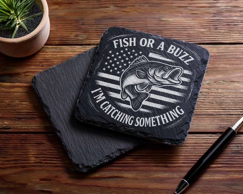May include: Two square black slate coasters. One coaster has the text "FISH OR A BUZZ I'M CATCHING SOMETHING" with a fish graphic and an American flag design. The other coaster is plain. A pen is on the wooden surface.