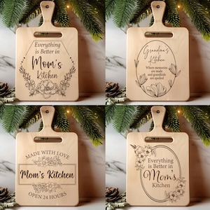 Puede incluir: Cuatro tablas de cortar de madera con asas, cada una grabada con un mensaje diferente con temática de cocina y diseños florales. Los mensajes incluyen "Everything is Better in Mom's Kitchen", "Grandma's Kitchen" y "Made with Love Mom's Kitchen Open 24 Hours."