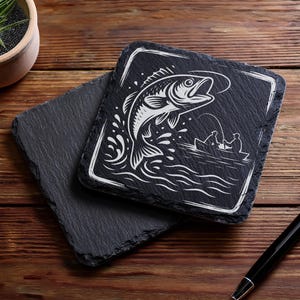 May include: Two square black slate coasters. One coaster features a white illustration of a fish jumping out of water with a fishing boat in the background. The other coaster is plain black. A pen and a potted plant are also in the image.