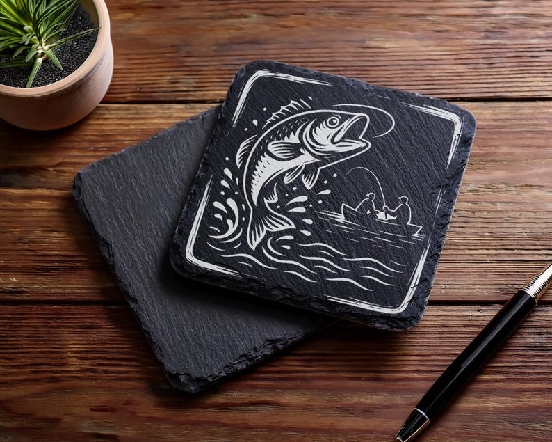 May include: Two square black slate coasters. One coaster features a white illustration of a fish jumping out of water with a fishing boat in the background. The other coaster is plain black. A pen and a potted plant are also in the image.