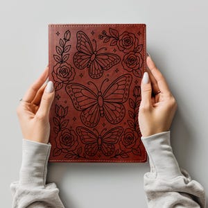 May include: A rectangular brown notebook featuring a black line art design of three butterflies surrounded by roses, leaves, and small star shapes. The cover has a smooth, leather-like texture with visible stitching along the edges of the front cover.