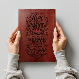 May include: A rectangular, dark brown leather-textured journal featuring black laser-engraved text that reads: Hope DOES NOT PUT US TO Shame, BECAUSE GOD'S LOVE HAS BEEN POURED OUT IN OUR HEARTS THROUGH THE Holy Spirit. ROMANS 5:5.