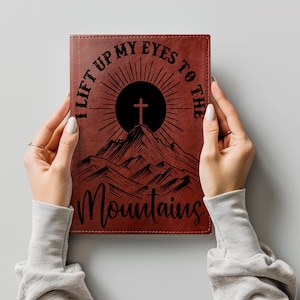 May include: A brown leather-textured journal featuring a black graphic of a mountain range with a sun and a cross at the peak. The text "I LIFT UP MY EYES TO THE" is printed in black above the graphic, and "Mountains" is printed below in script.