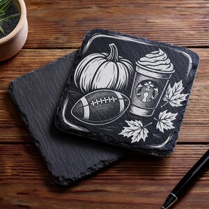 May include: Black slate coasters with white etched designs. One coaster features a pumpkin, football, coffee cup, and maple leaves. The other coaster is plain. The coasters are on a wooden surface.
