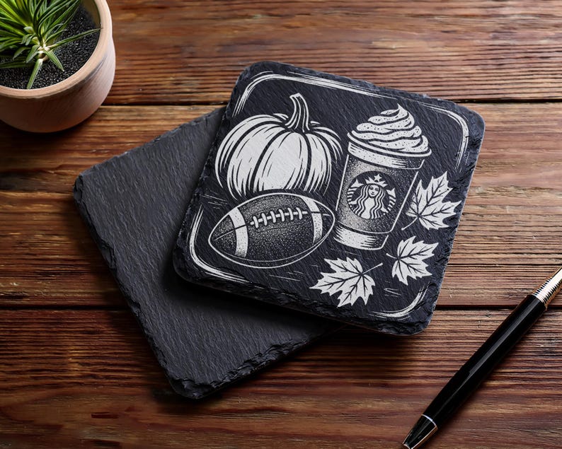 May include: Black slate coasters with white etched designs. One coaster features a pumpkin, football, coffee cup, and maple leaves. The other coaster is plain. The coasters are on a wooden surface.
