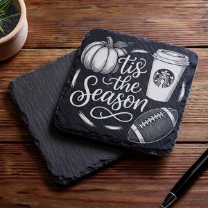 May include: Two square black slate coasters. One coaster has a pumpkin, coffee cup, and football design with the text "'tis the Season" in white. The other coaster is plain. A pen rests on the wooden surface.